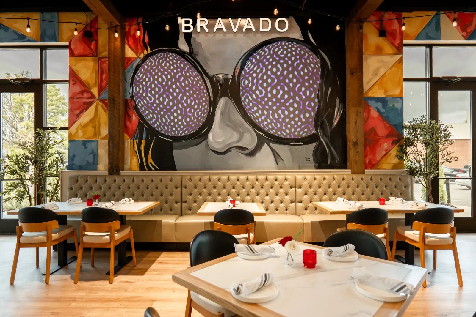 The Sunroom at Bravado: tufted banquette and tables beneath the painted sunglasses mural and bistro lights
